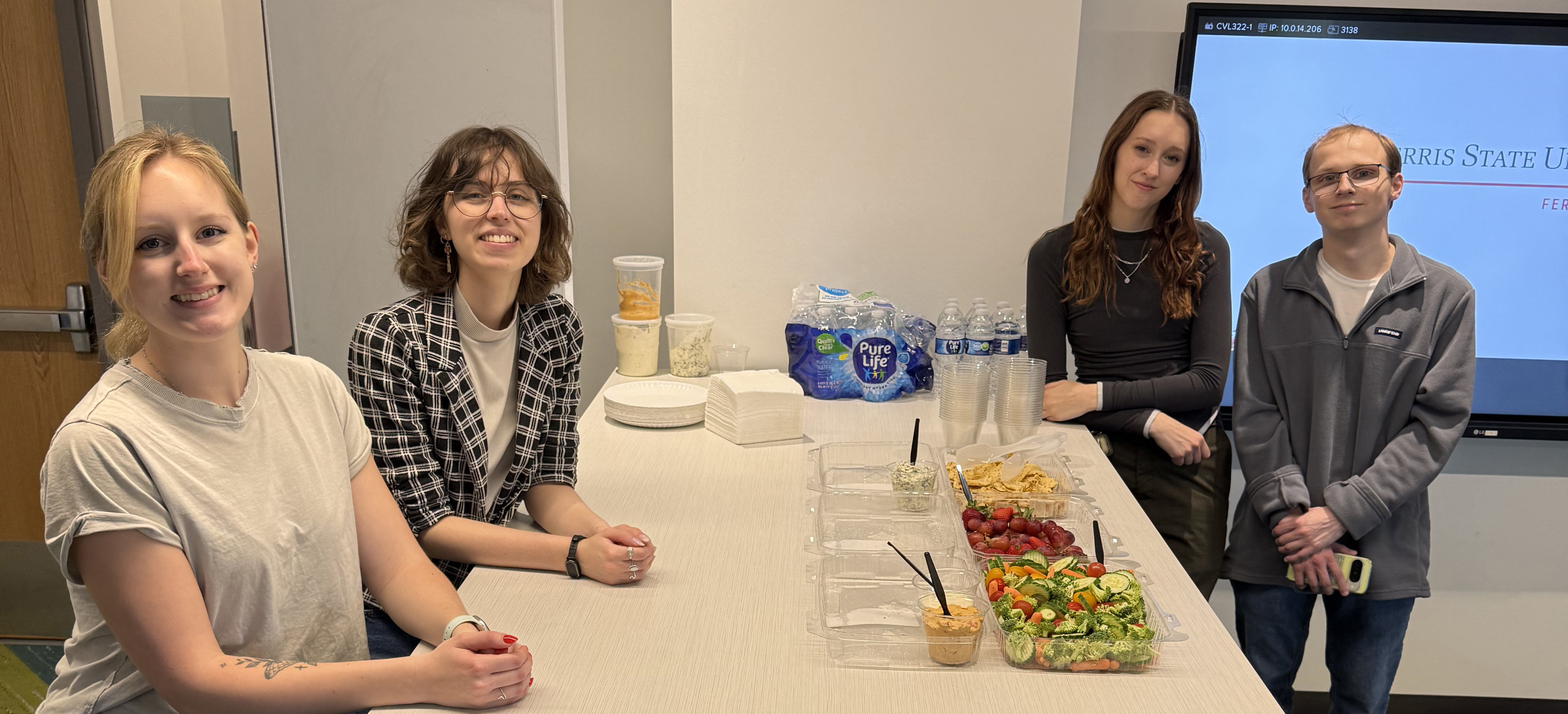 Emma Eling, Hailey Nye, Abby Keown, and Blake Flannery smiling at the camera by a table featuring small snacks
